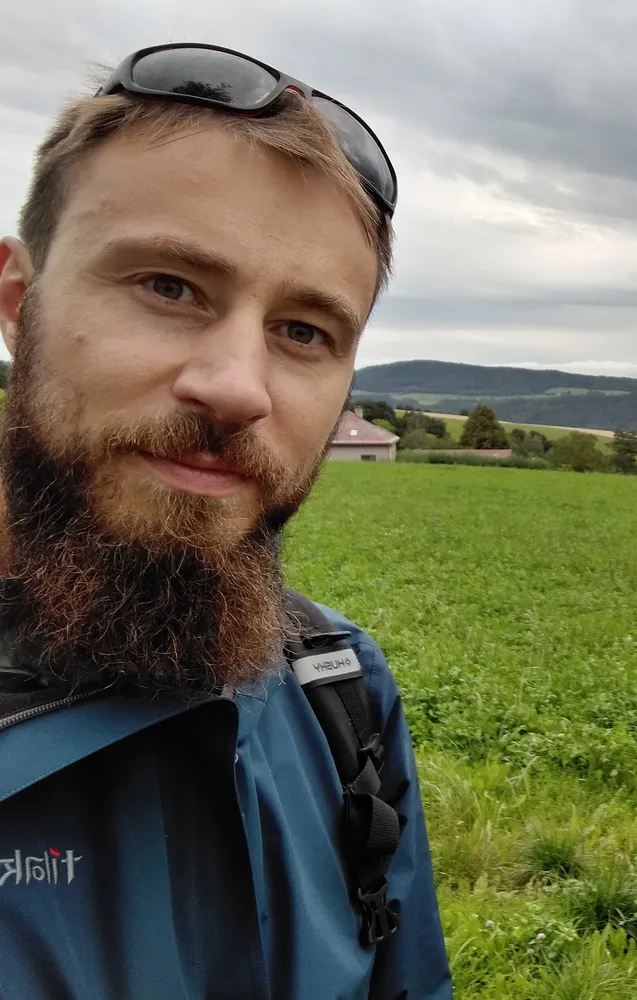 Autor blogu upřeně kouká do záběru, za ním je zelené pole a vesnice