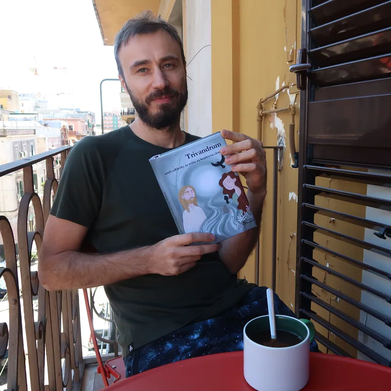 Autor blogu sedí na židli na terase. V rukou má svůj román Trivandrum.