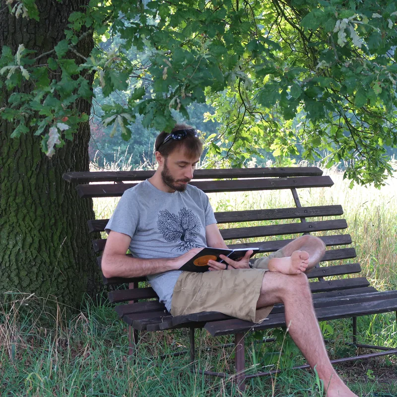 Autor blogu sedí pod stromem na lavičce a píše do notesu.