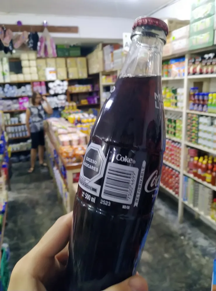 Skleněná lahev Coca-Coly s upozorněním 'exceso azucares' (nadměrný obsah cukru) ve španělštině, focená v typickém mexickém malém obchodě s regály plnými zboží v pozadí.