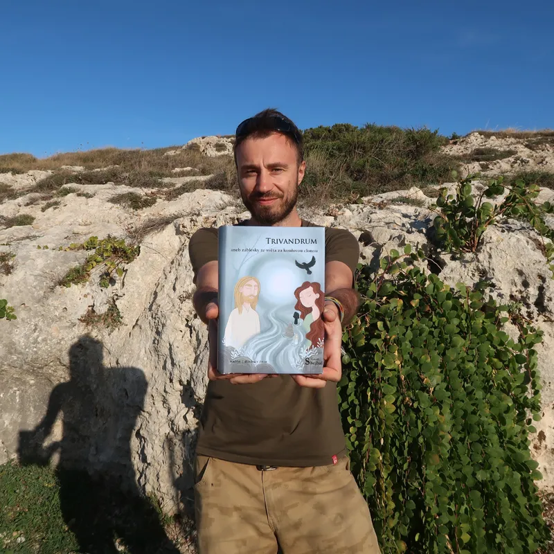 Autor románu Trivandrum drží před sebou knížku, v pozadí kameny a keř